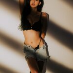 Sensual young slim female in elegant lingerie and denim shorts leaning on wall with arm above head and looking at camera in sunlight